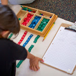 This 1st year student is practicing addition with carrying with the stamp game into the thousands. In the Common Core curriculum, by contrast, dynamic addition beyond 1,000 isn't introduced until 3rd grade.