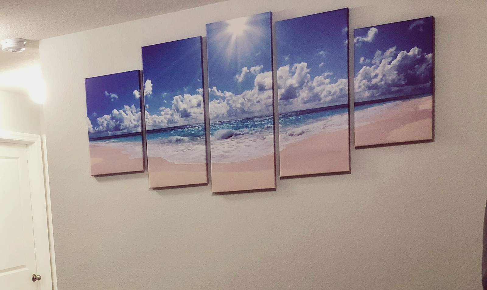 Five canvases displaying a painting of a beach on a off-white wall. 