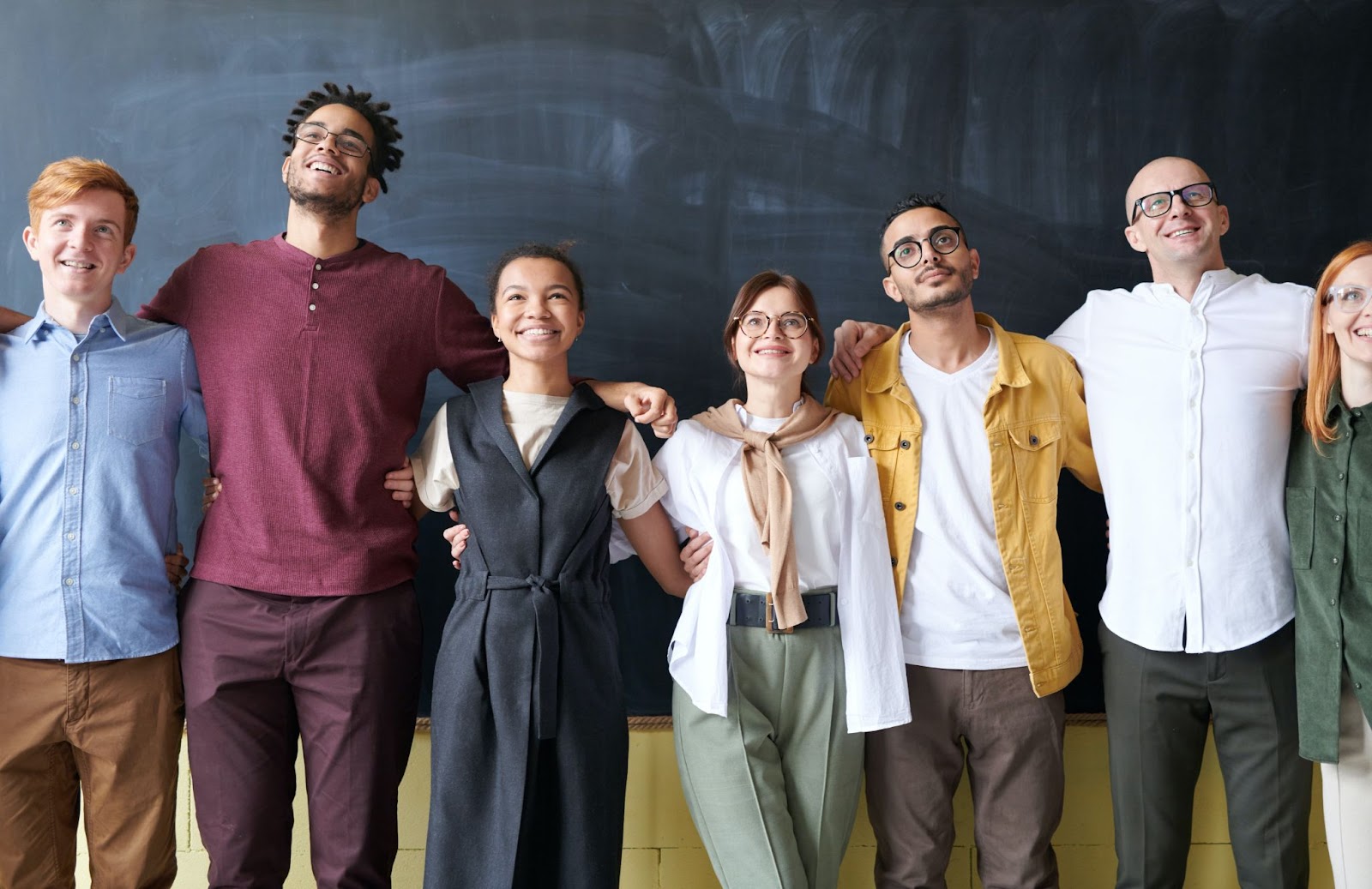 A imagem mostra sete pessoas que se abraçam lateralmente com expressões sorridentes. São quatro homens e três mulheres, com diferentes estilos e figurinos. Eles estão em frente a uma lousa negra. 