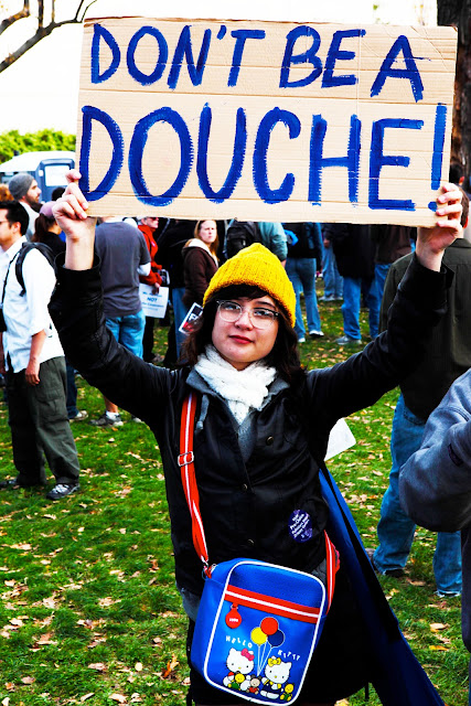 A hipster with a Hello Kitty purse and a bright yellow beanie holding a sign that says, "Don't Be A Douche".