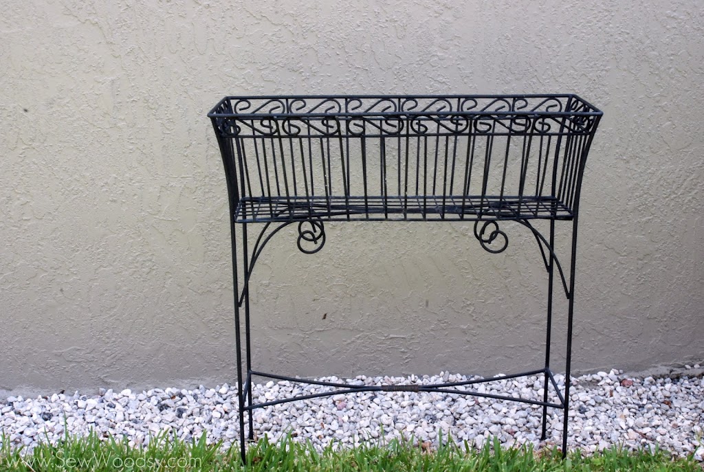 Black wire raised plant stand against a grey wall resting on rocks and grass.