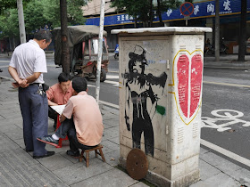 two men playing xiangqi next to an electric box with a drawing of a woman
