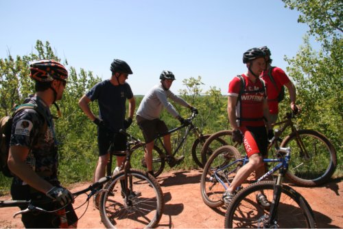 A group of cyclists, sitting on their Surly bikes, on a red dirt mound, with trees and blue sky behind them