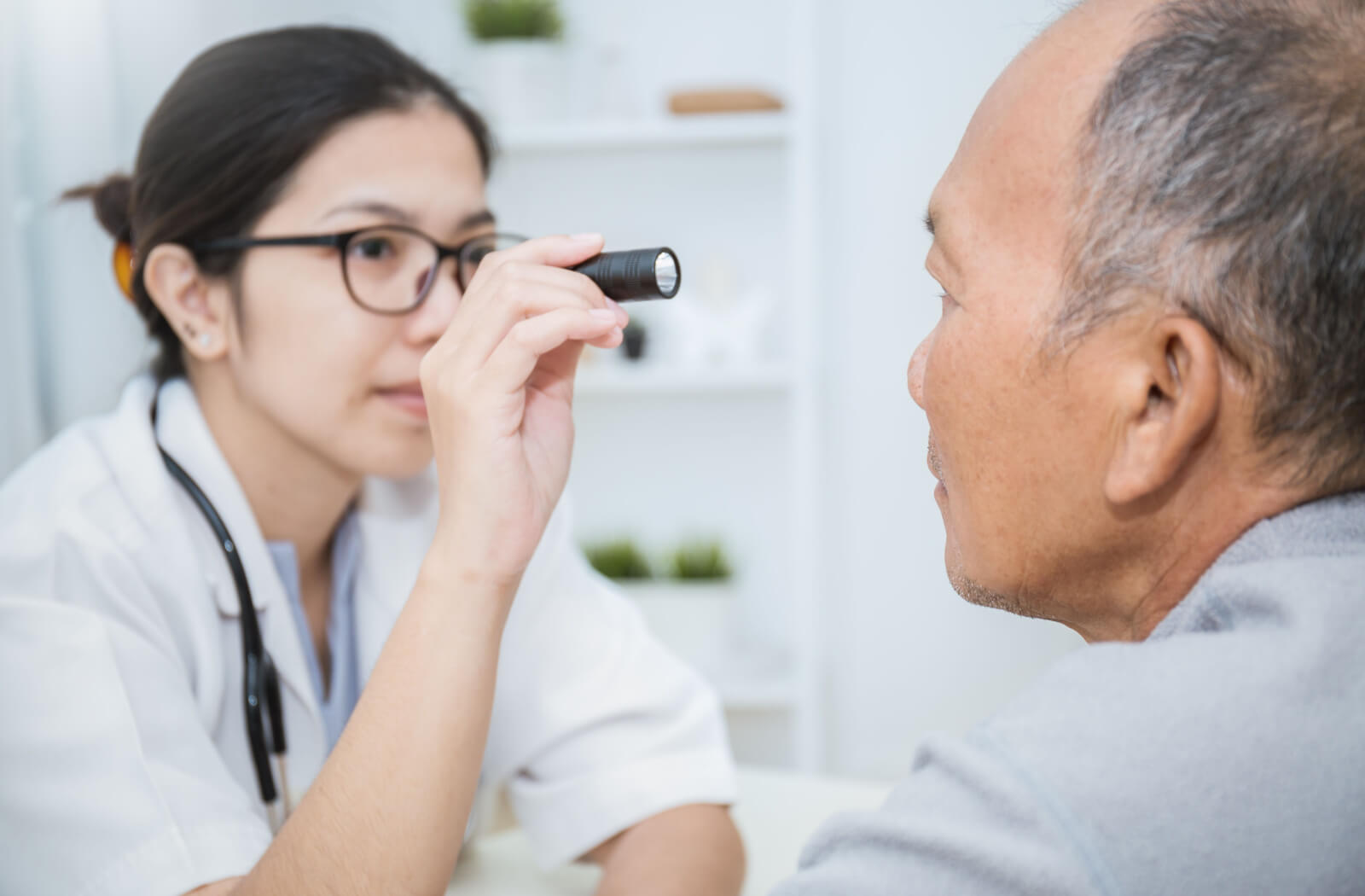 A senior man facing his optometrist as she shines a small flashlight into his eye.
