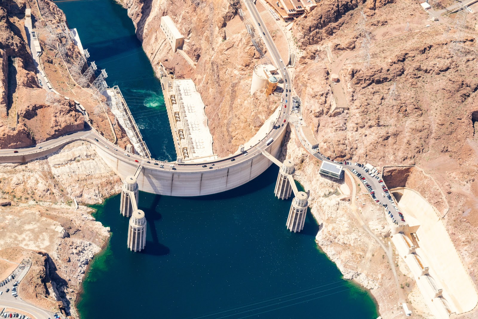 Arial view of the Hoover Dam with brown rock and desert on each side and blue-green water on both sides of the dam. 