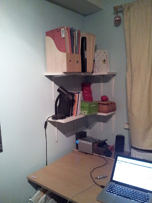 Same desk, less stuff on the desk, white shelves fixed to the walls above the back left corner of the desk