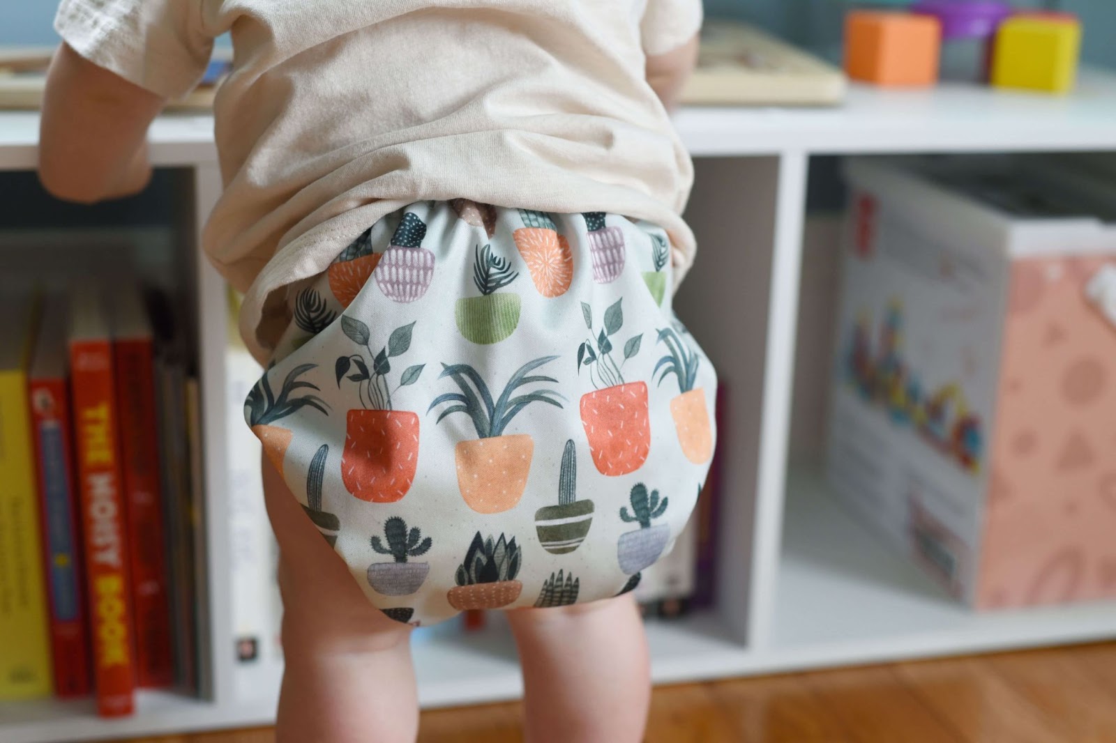 Making Cloth Diapering Cute Again With Henry and Howie–The image shows the back of a baby standing in front of a bookshelf. The baby is wearing a Henry and Howie cloth diaper that has a light blue background with a cartoon design of colorful potted plants. 