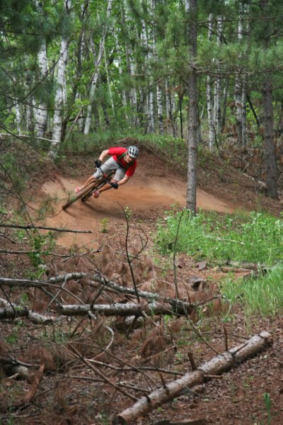 Front view of a cyclist riding a Surly bike, rounding a berm from right to left, on a dirt trail in the forest