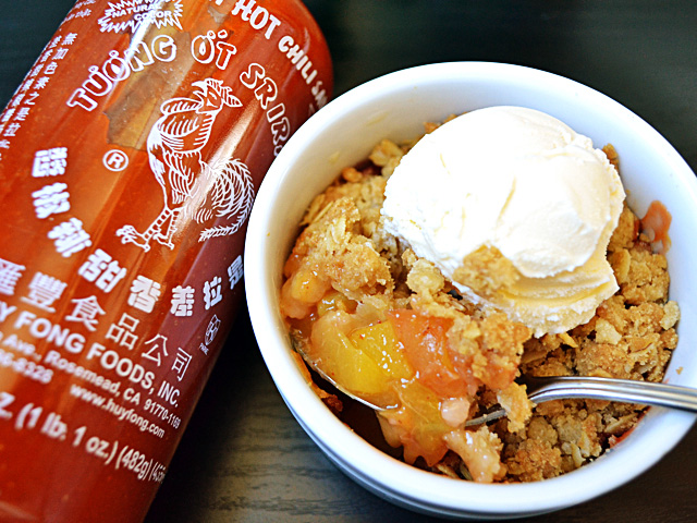 Sriracha Peach Crisp in bowl topped with scoop of ice cream, with bottle of Sriracha on the side 