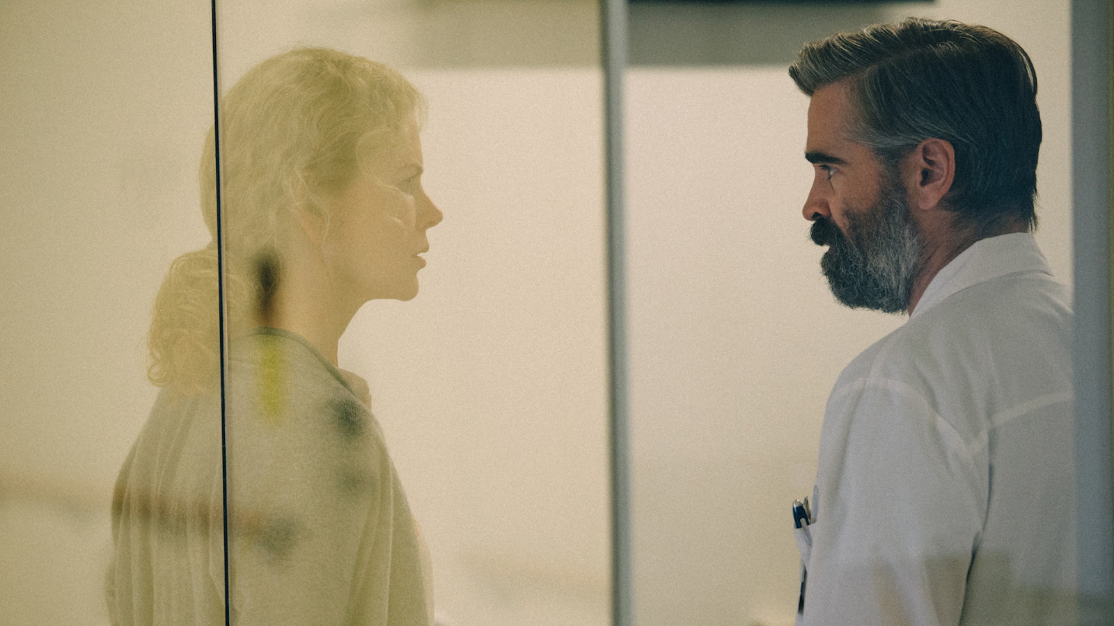 A middle-aged man wearing a white coat looks at a woman around the same age. A pane of glass separates them.