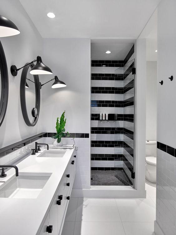 banheiro com azulejos brancos e faixas de azulejos pretas, área do box com piso de pastilhas pretas, piso branco no demais, armário branco com puxadores pretos, torneiras e luminárias pretas e espelho redondo com moldura preta.