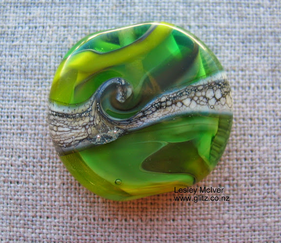 This is a bead showing the flow of the glass – like water – with the swirl of the land meeting the sea. Multiple greens and silvered ivory glass. Artist Lesley McIver.