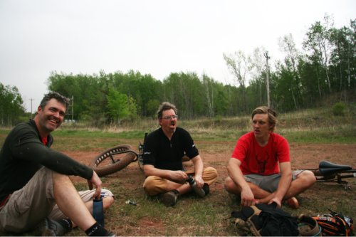 Front view of 3 cyclists, sitting on a dirt & grass field, with their Surly bikes laying next to them