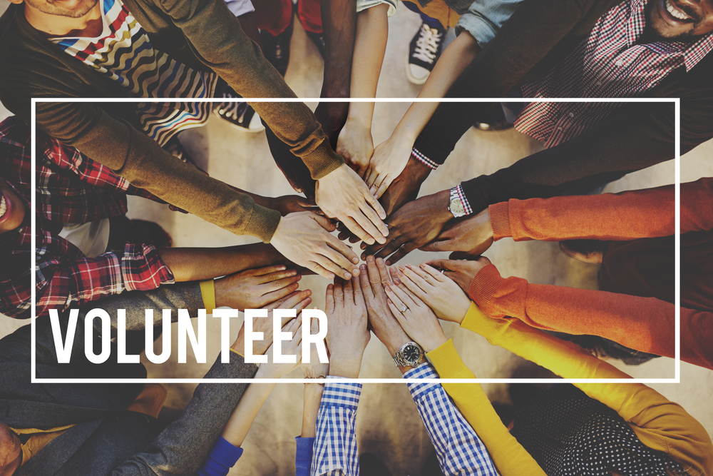A group of people standing in a circle holding a hand out to the center with the word Volunteer superimposed over the photo.