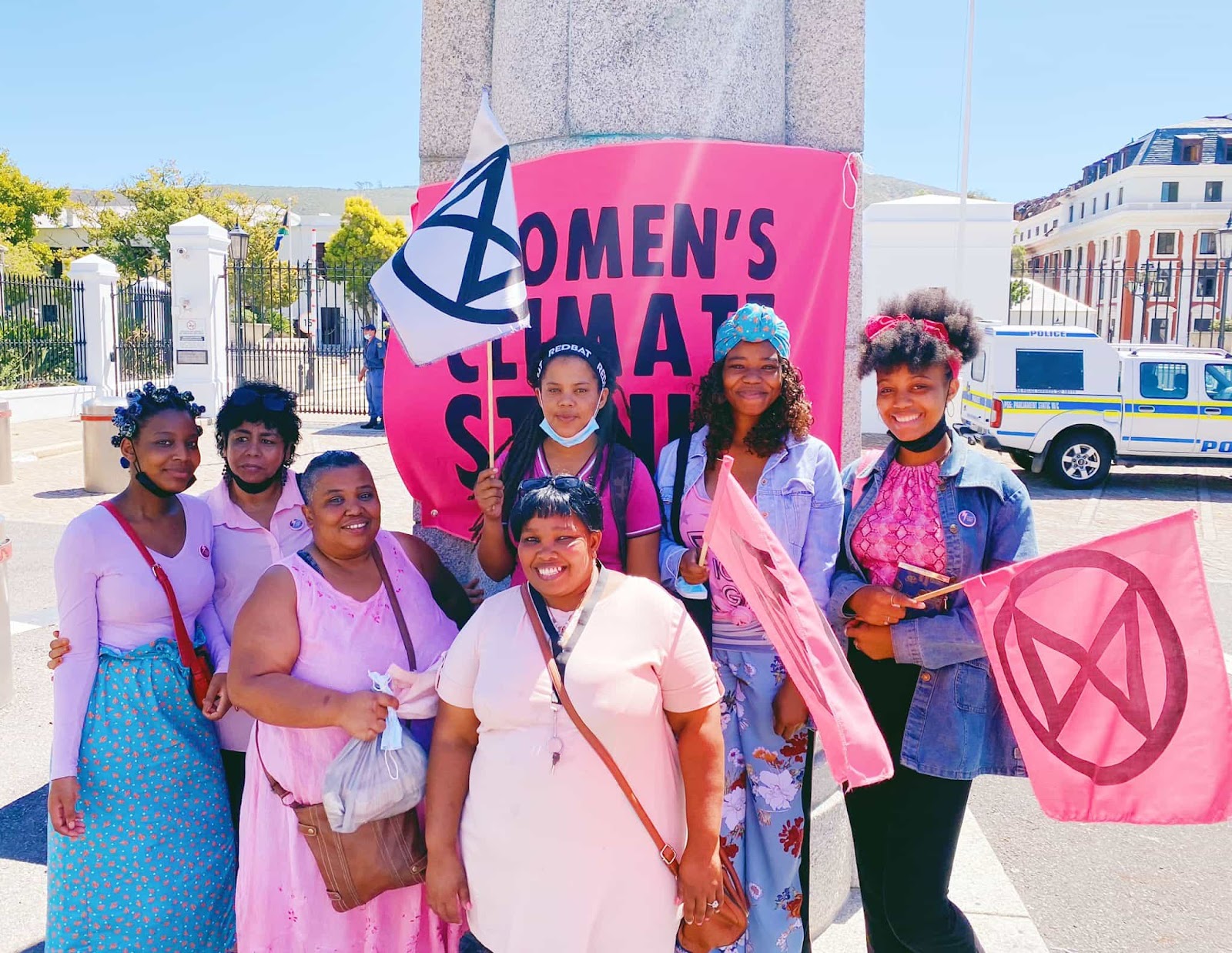 A group of women wearing pinks and purples and holding XR flags smile in front of a banner saying women's climate strike