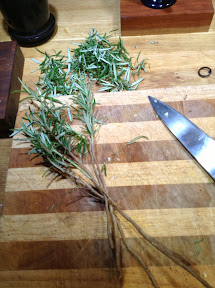 Four rosemary sticks and a pile of rosemary leaves on a chopping board, knife next to them