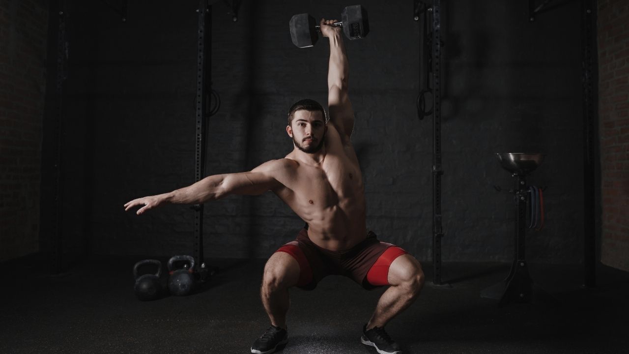 A man performs single-arm overhead squats with a dumbbell in his working hand.