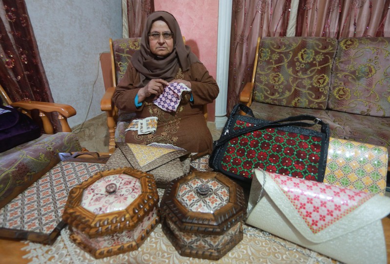 Older woman sits in chair and works on embroidery panel while surrounded by boxes and handbags decorated with embroidery