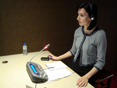 A female-presenting individual stands in front of a table-mounted microphone wearing a headset. There are notes on the table in front of her. 