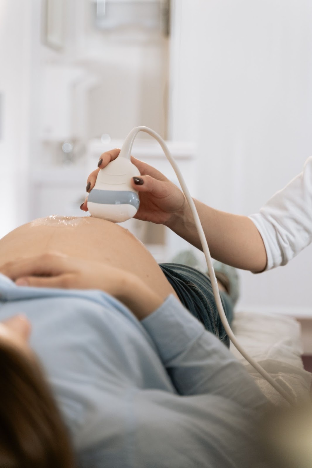 A imagem mostra um exame de ultrassom sendo realizado em uma mulher grávida. A gestante tem cabelos morenos, veste uma camisa social azul bebê com calça jeans e está deitada em uma maca cirúrgica, enquanto uma médica loira com unhas pintadas de marrom realiza o procedimento.