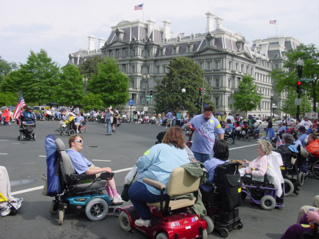 Many people using wheelchairs are in the street with a Victorian four-story building in the background. 