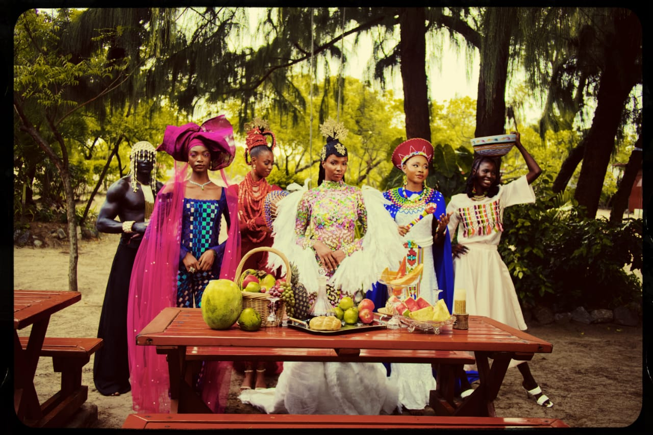 Models dressed in African traditional attire behind a table laced with fruits.