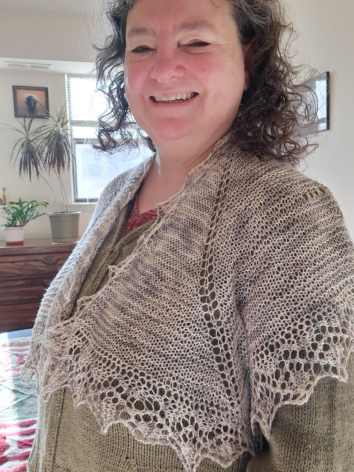 White woman with brown curly hair smiling wearing a white-grey shawl over her shoulders. There are plants and pictures in the background. 