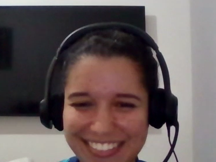 Foto de Dayana, uma mulher branca de cabelos escuros e presos. Na imagem ela está sorrindo e usando fones de ouvido extra auricular.