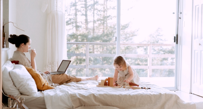 a woman working from home in bed as her child plays nearby
