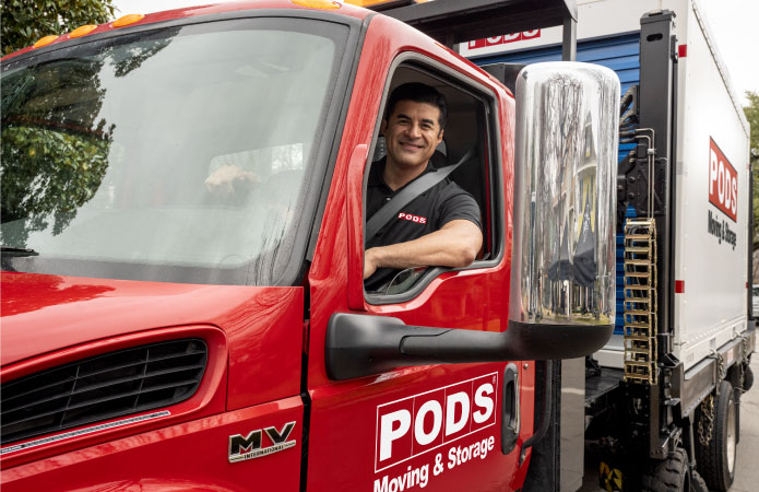 A smiling PODS driver sitting in his red PODS truck while transporting a PODS portable moving container.