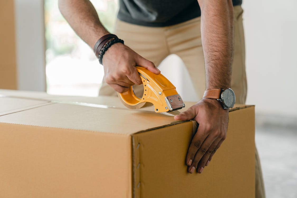 Crop faceless young male with wristwatch using adhesive tape while preparing cardboard box for transportation