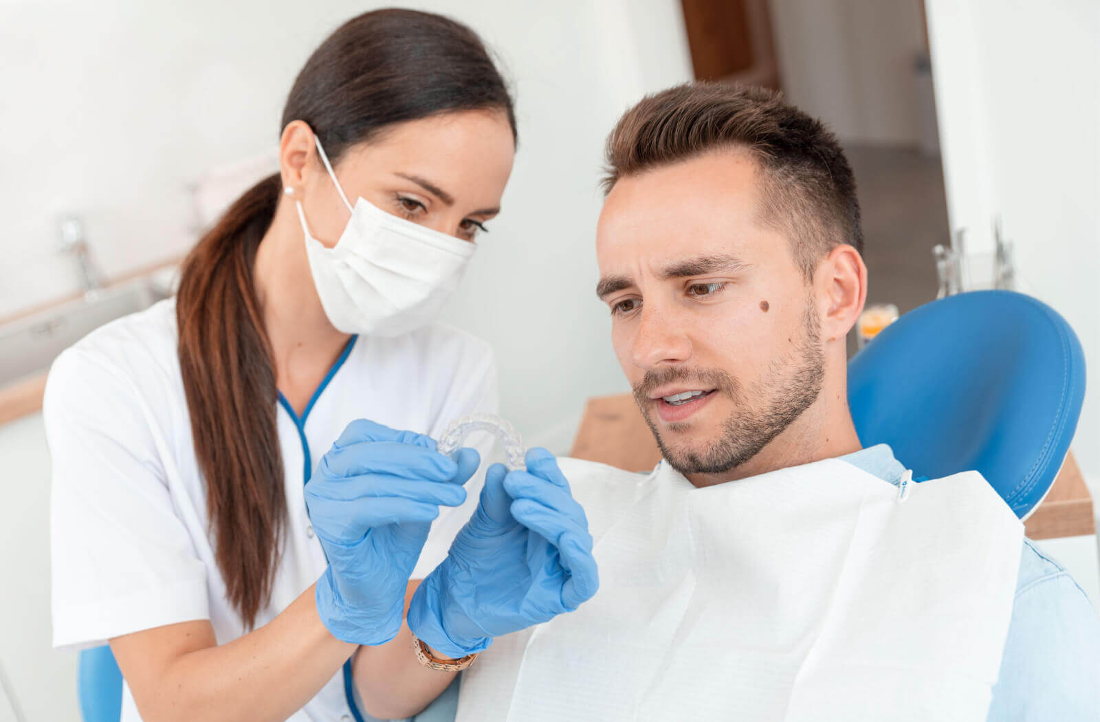 A female dentist in her white uniform with blue latex gloves on her hands is holding a new Invisalign brace while demonstrating it to her male patient that is sitting on a dental chair.