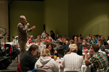 A male-presenting individual speaks into a microphone he's holding while standing in front of a large group of people, all seated at tables. The people appear to be eating a meal. 