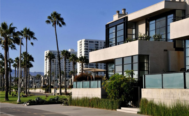 A modern Los Angeles apartment building with concrete and glass elements. There’s decorative landscaping in the front of the building, a number of tall palm trees nearby, and other larger apartment buildings in the background.