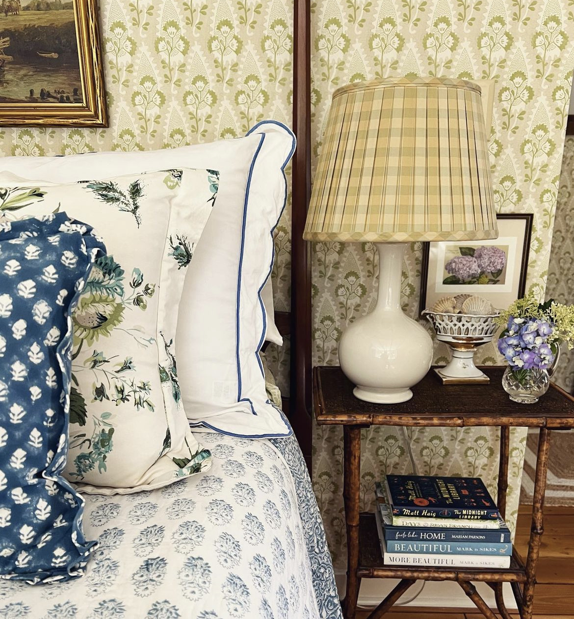 The bed is flanked by a vintage bamboo style table decorated with a lamp, books, and some bric-a-brac.