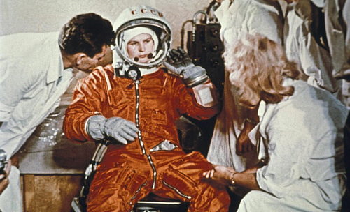 Valentina Tereshkova sentada, con el traje, guantes y casco de astronauta, hablando con dos personas.