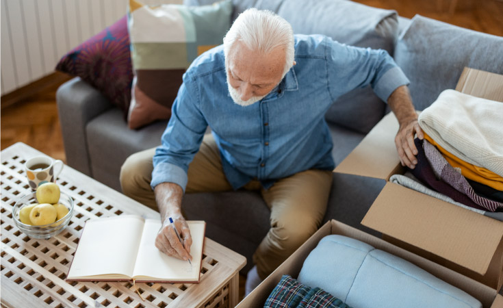 A mature man is making a note as he packs moving boxes with blankets and other linen.