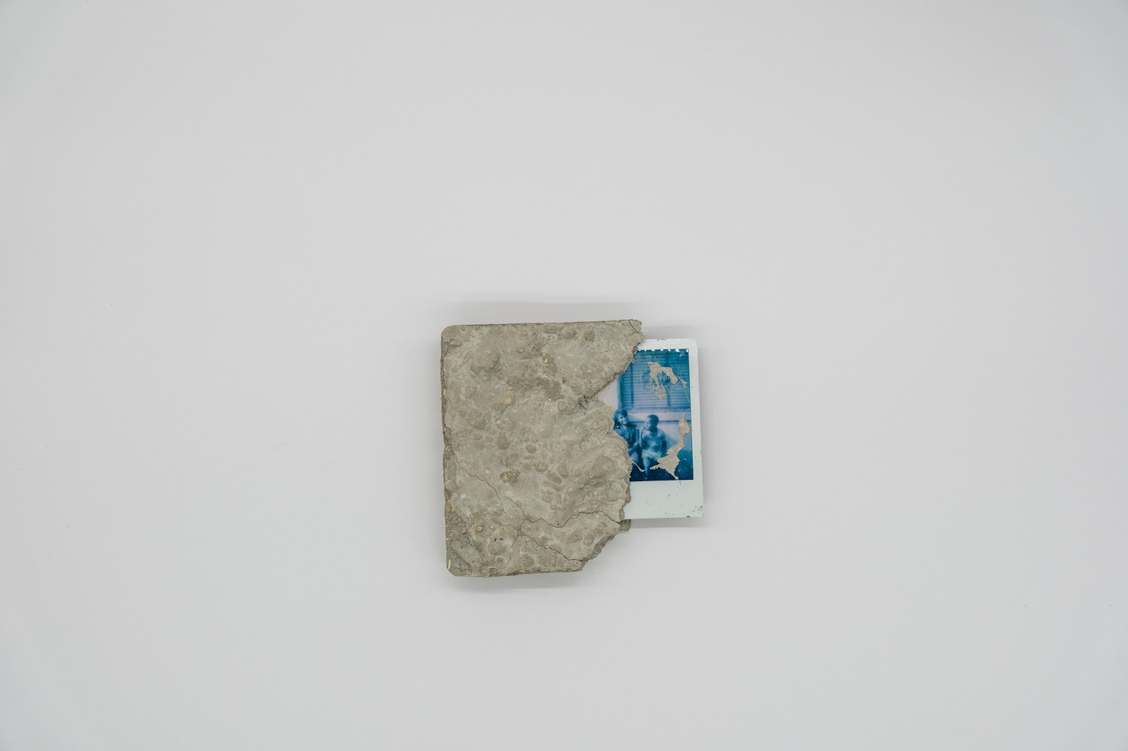 Image: A detailed view of the 2002 work, In Remembrance of Us. The work is a square of concrete with parts chipped away along the right edge. A blue-hued Polaroid photo of two people is encased by the concrete along the right edge. Photo courtesy of Sibyl Gallery.