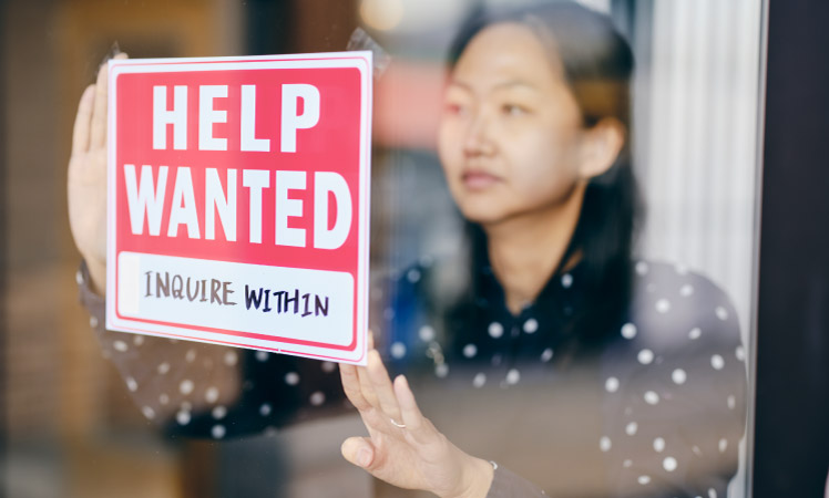 A woman tapes a sign in the window of her business that reads, “HELP WANTED. INQUIRE WITHIN.”