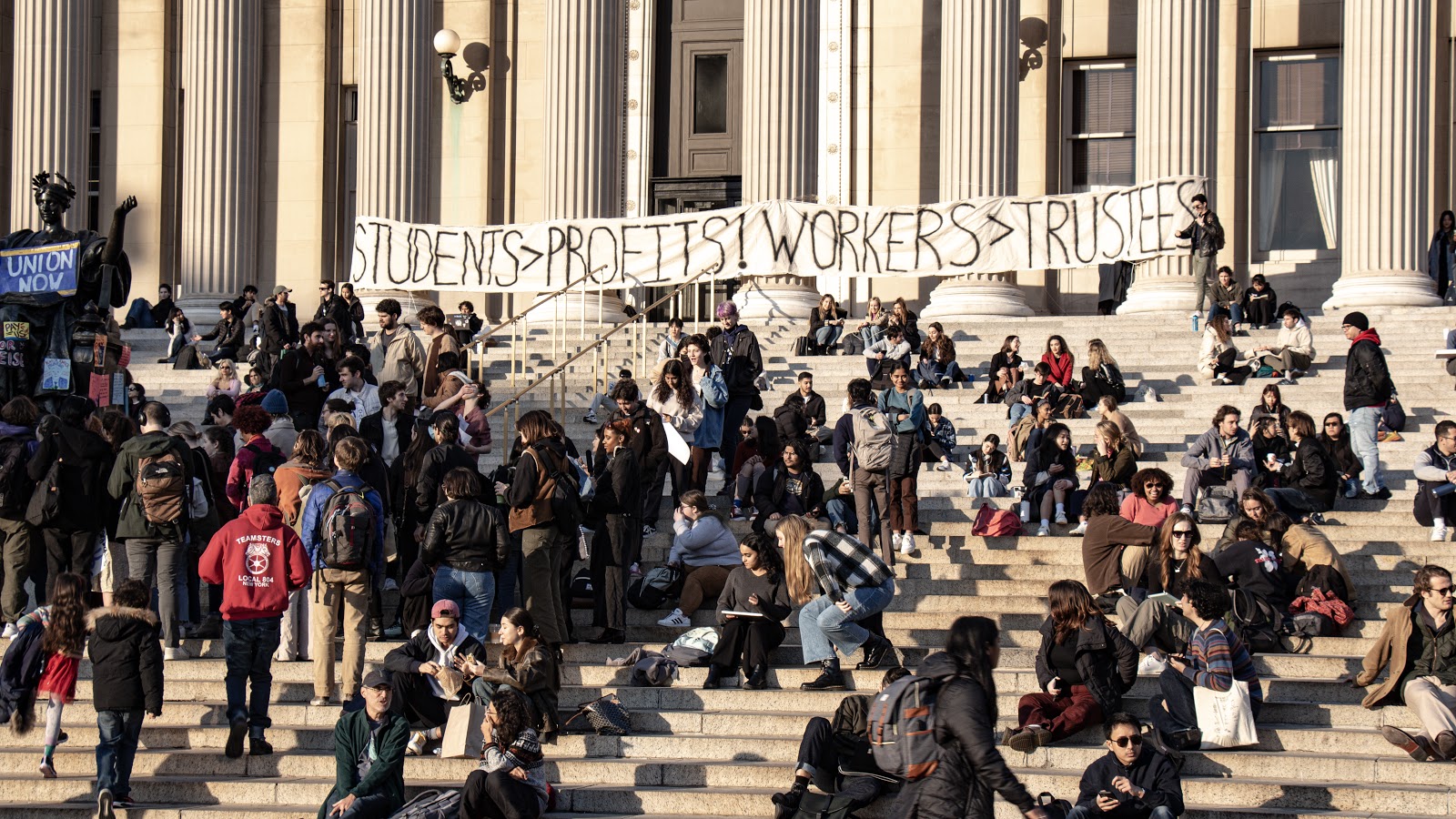 A large crowd of students is pictured on the steps of a library that is supported by large white pillars. Tied to the pillars is a large, canvas banner that says, in all capital letters, “Students > Profits! Workers > Trustees!” in black letters.
