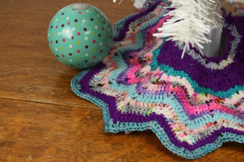 a multicoloured crochet star tree skirt round the bottom of a tiny plastic christmas tree and a large green polka dot bauble hanging on one side