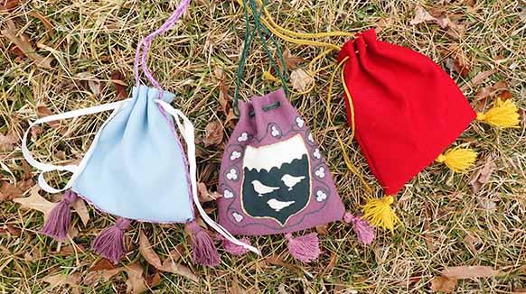 Three drawstring bags on the grass. On the left is a sky blue drawstring bag with three purple tassels, a purple belt cord, and white fabric drawstrings. The middle bag is a purple drawstring bag with lighter purple tassels, and a forest green cord. On the front is a shield with a white space at the top and black at the bottom. The black space has regularly occurring humps giving the white a cloud like appearance and there are three white birds on the black. Around the shield is a wreath of white flowers on a green vine, each with three petals. On the right hand side is the red and gold bag made for this article.
