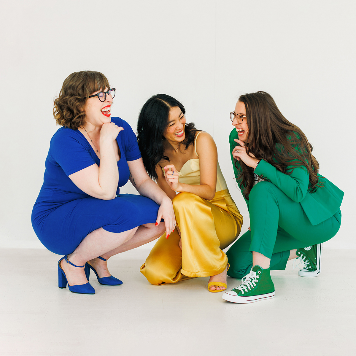 Photo of the Rendezvous Creative team crouched in laughter in a white room. Megan, in a blue dress, blue heels, and red lipstick laughs with her hand under her chin. Nicole, in yellow pants, leans back on her hand while laughing at Latasha on the right, who is dressed in a green suit and green Converse shoes.