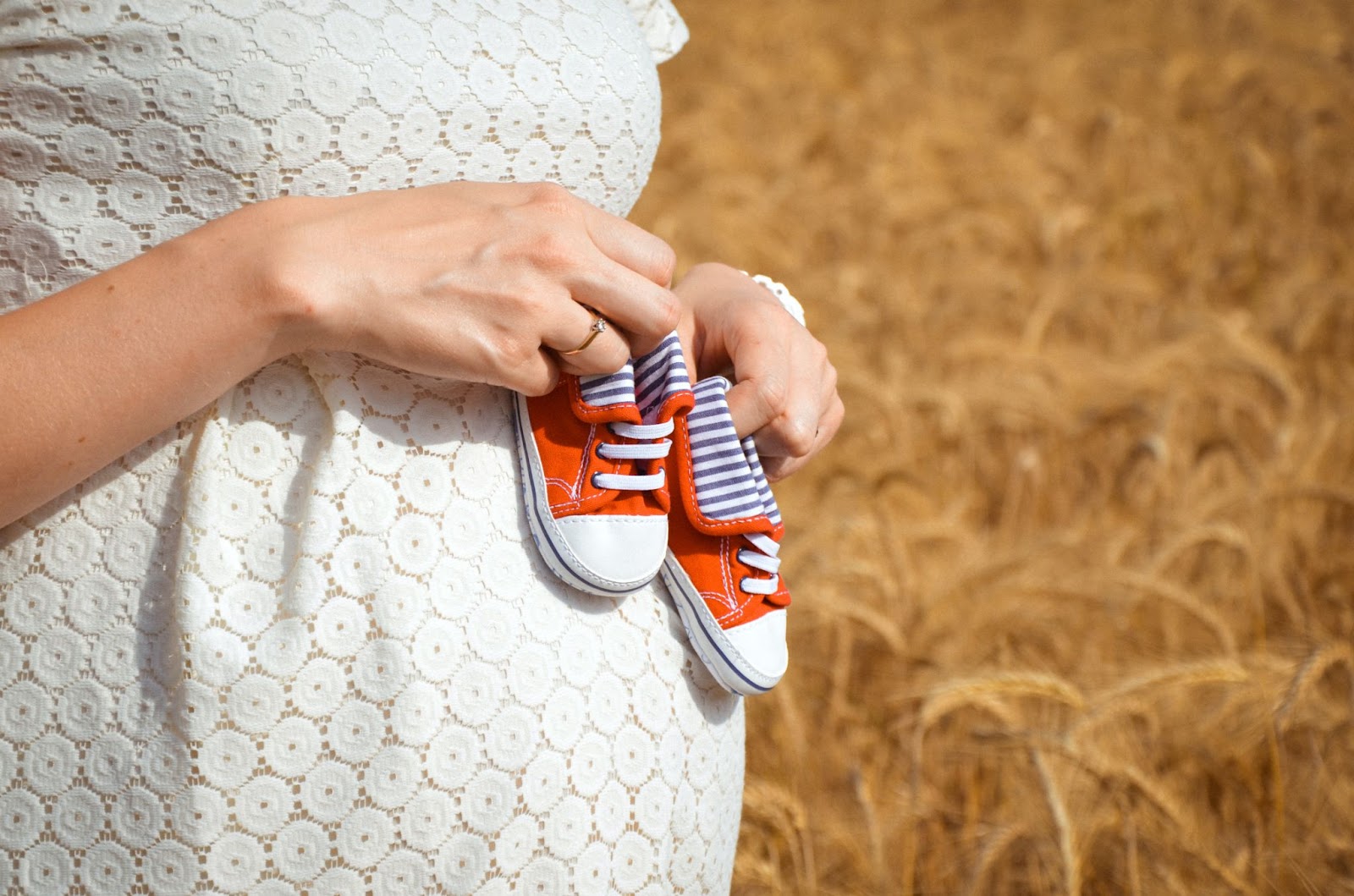 an expecting mother in a white dress, holding two small converse-style baby sneakers