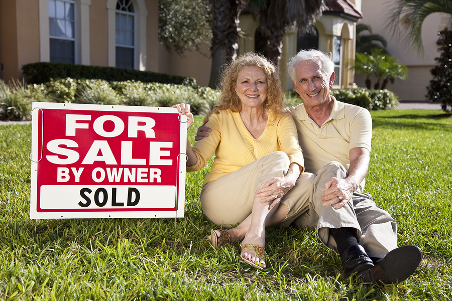 Mature couple on lawn of their FSBO for sale by owner home smiling with a sold sign