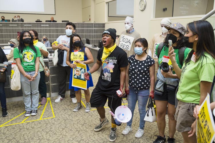 A young activist shouts out to the conference room. Other young activists stand beside him. Behind older rebels dressed in white banages with signs of horrors to come watch