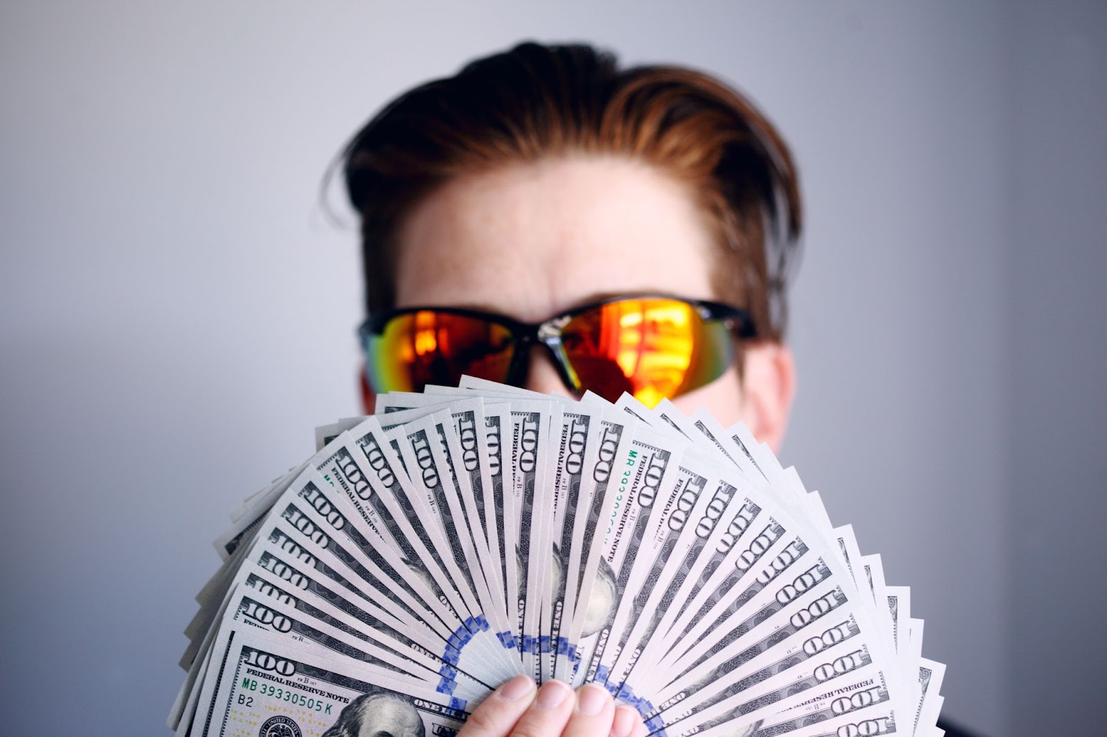 Picture of a man wearing sunglasses, holding several one hundred dollar bills in a fan shape.
