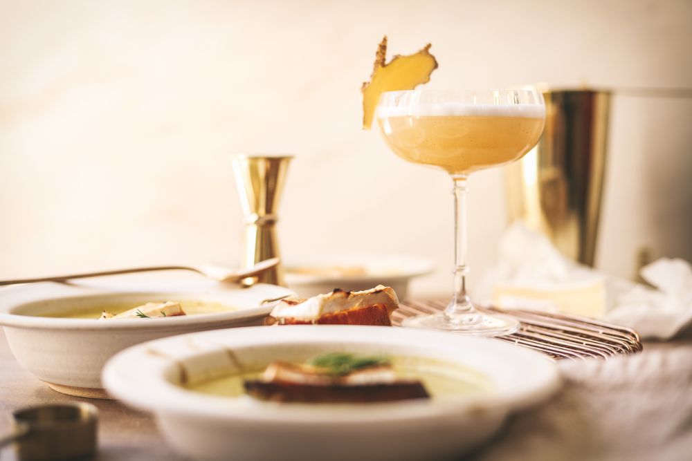 A ginger non-alcoholic cocktail in a coup glass, surrounded by soup. 