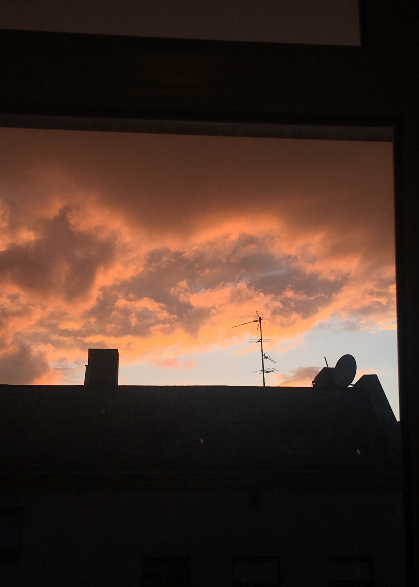 A sunset over a rooftop from a window.  The clouds are grey, orange, purple and pink with a sliver of blue sky.  A chimney, satellite dish, and antennae are silhouetted.  