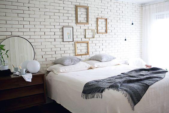 Quarto com cama de casal, parede da cabeceira com azulejo subway tiles branco, quadros com molduras douradas, criado mudo de madeira escura e espelho redondo.